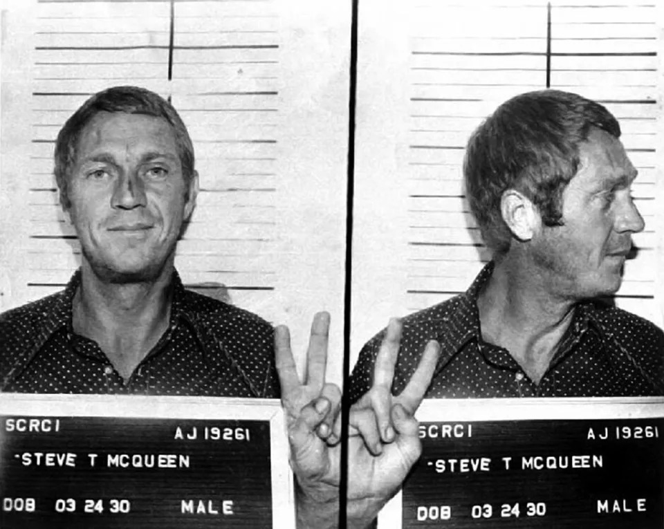 Mugshot of Steve McQueen with front and side profiles, holding a name placard; he gestures a peace sign with his hand